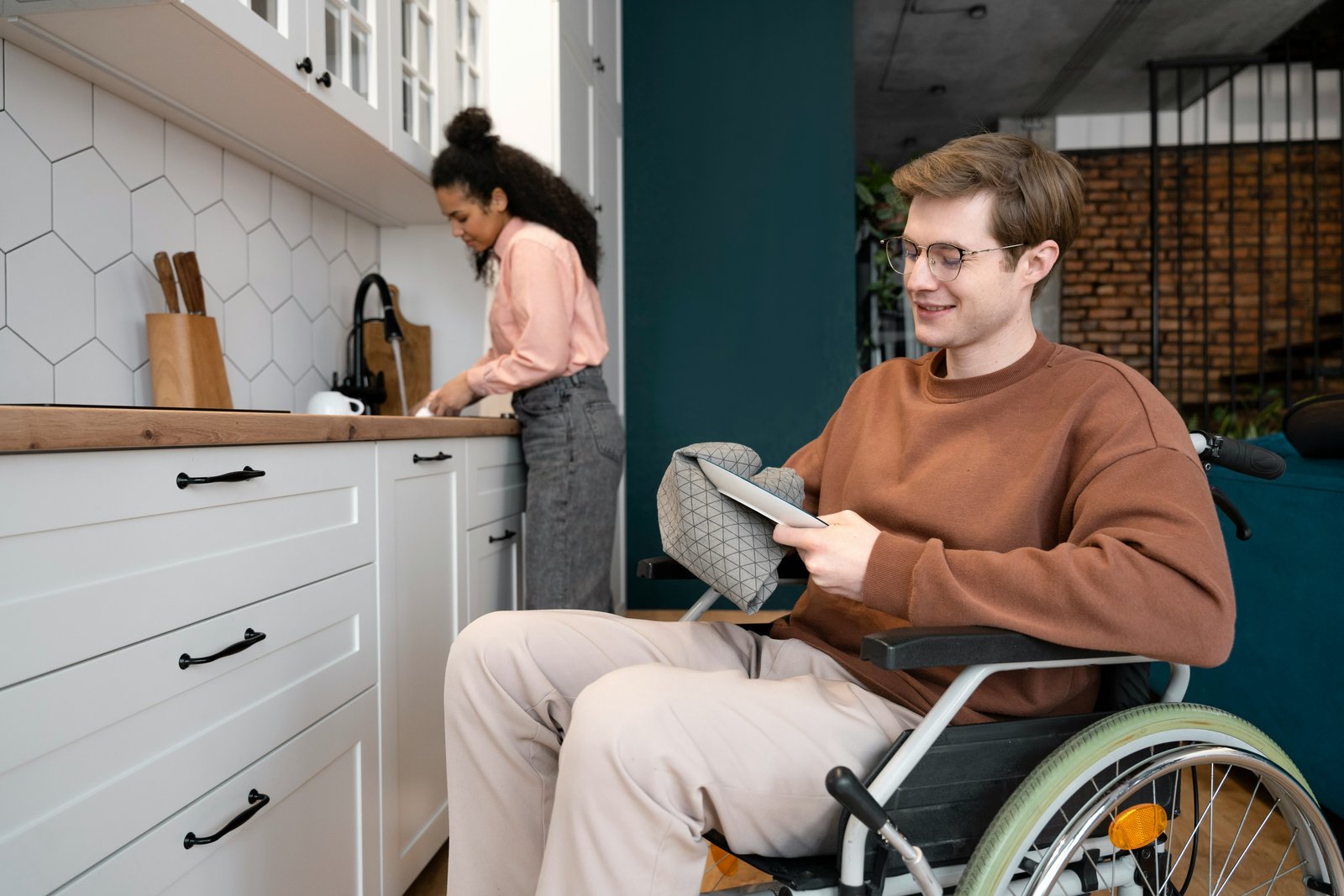 A man using a wheelchair using a tablet in a kitchen while a housemate washes dishes nearby.